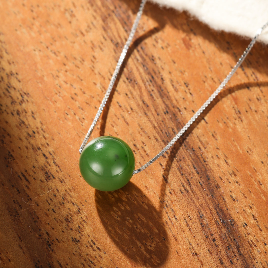 Close-up of a smooth, round green jade pendant on a delicate silver chain, resting on a warm wooden surface.