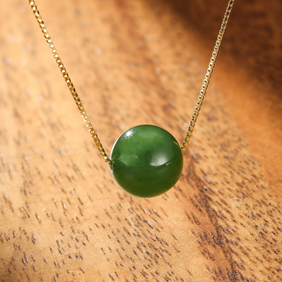 A single, smooth, round, deep green jade pendant hangs on a delicate gold chain, set against a warm, textured wooden background.