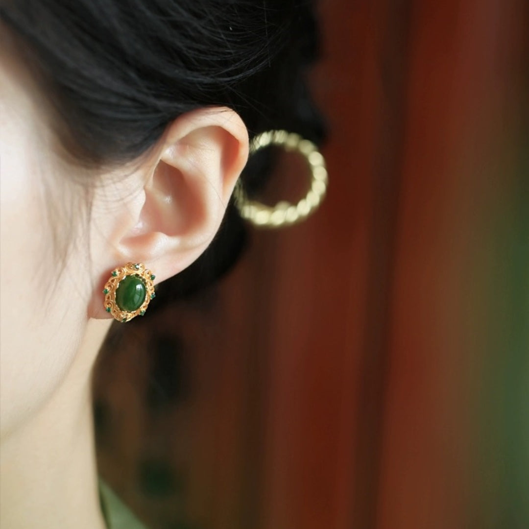 Close-up of a woman's ear wearing Charmed Green Jade Cloud Stud Earrings, featuring a central oval jade stone surrounded by smaller green gems and intricate gold-plated metalwork.