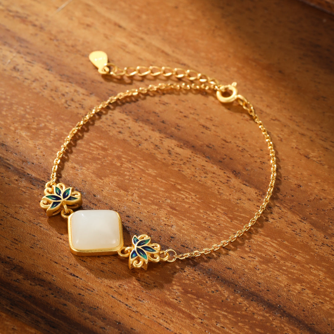 Gold-plated silver bracelet featuring a square white Hetian jade stone centerpiece, accented with blue enameled lotus flower charms on a wooden background.