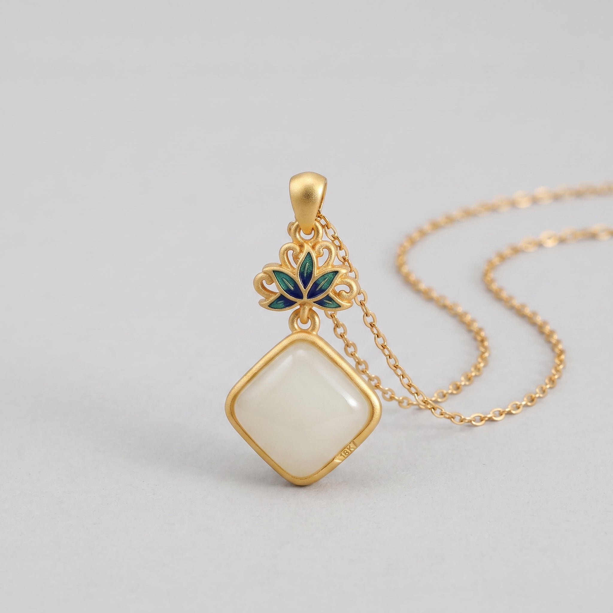 A delicate gold necklace featuring a rhombus-shaped, milky white Hetian jade pendant, adorned with an intricate lotus-inspired charm in green and blue enamel.