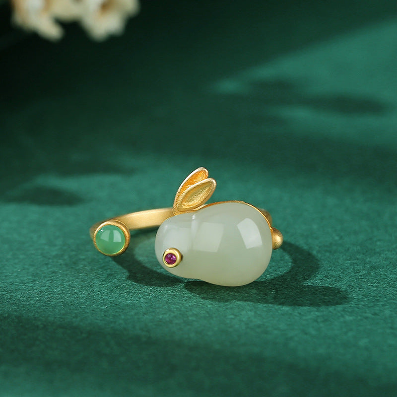 Moonglow Hetian Jade Rabbit Ring with gold-plated silver band and ruby eyes on a dark green velvet background.