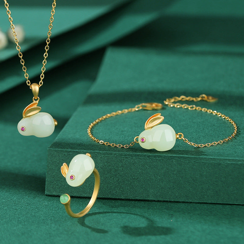 A matching set of Moonglow Hetian Jade Rabbit jewelry, including a pendant necklace, bracelet, and ring, each featuring a jade rabbit with ruby eyes on a delicate gold chain, displayed on a dark green velvet surface.