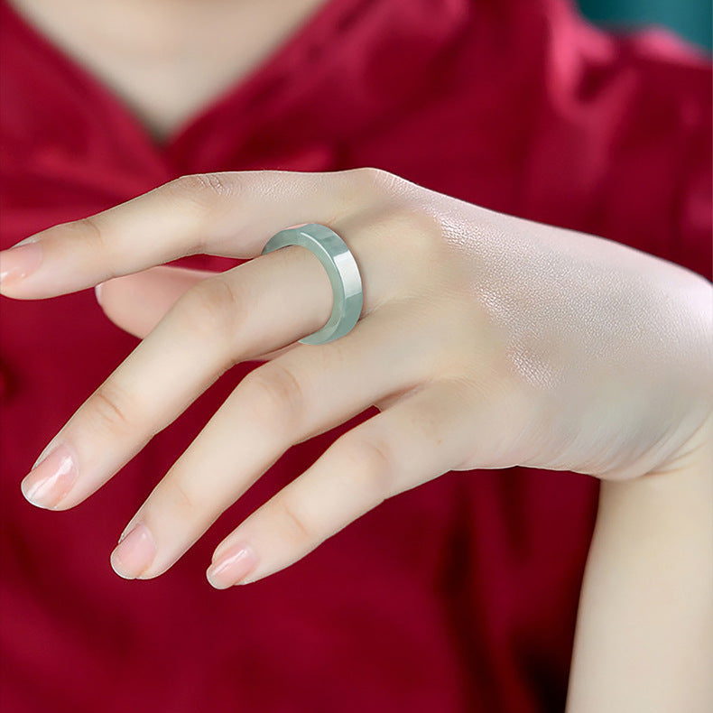 Close-up of a hand wearing a smooth, pale green ice jade band ring with a faceted exterior, faceted exterior, faceted edge, set against a rich red background.