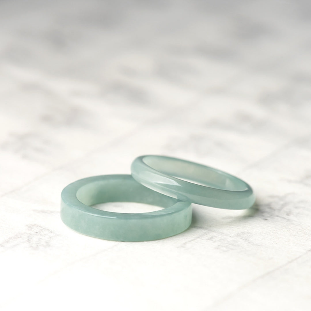 Pair of smooth, translucent ice jade band rings displayed on a light, textured surface. One ring is in focus in the foreground, and the other is slightly behind and to the right.