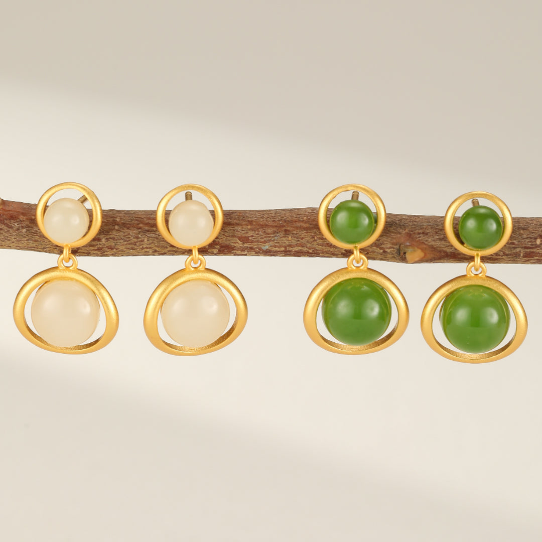 Solstice Hetian Jade Sun Drop Earrings in white and green jade with gold-plated silver frames, displayed on a wooden branch.