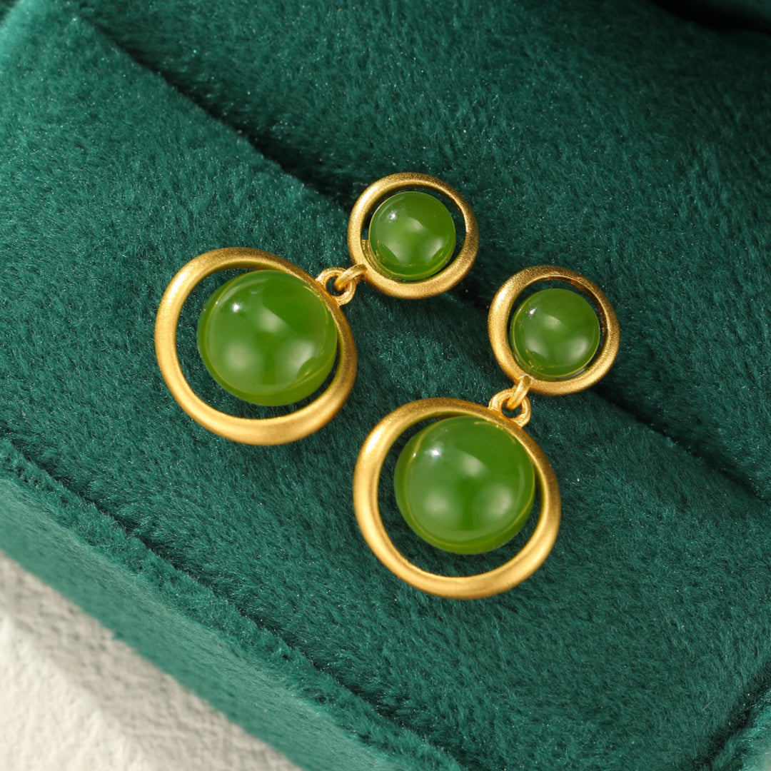 Solstice Hetian Jade Sun Drop Earrings featuring green jade spheres suspended within gold-plated silver circular frames, presented on a green velvet background.