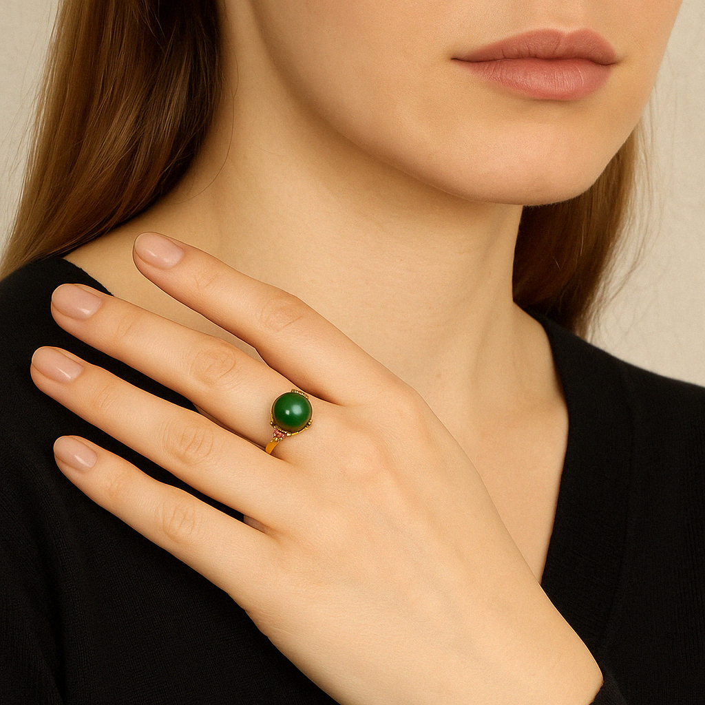A woman's hand wearing the Sovereign Green Jade Solitaire Ring on her ring finger. The ring features a large, round, deep green jade stone set in a gold-plated silver band, accented with small pink stones.