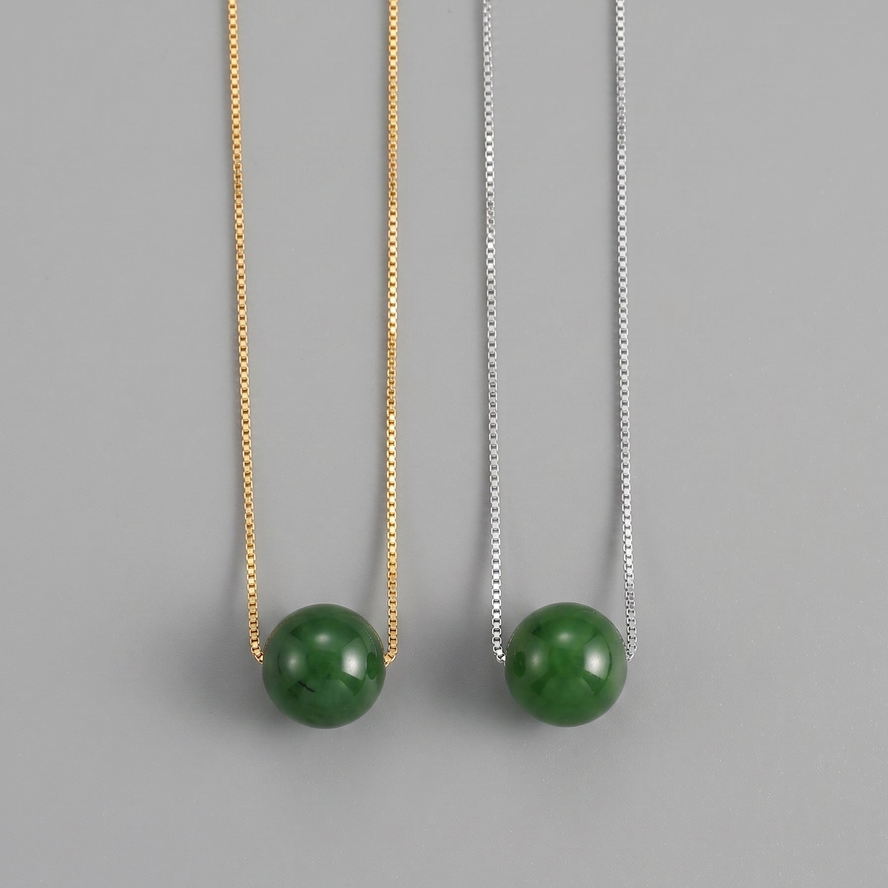 Two Acorn Green Jade Pendant Necklaces, one on a gold chain and one on a silver chain, against a grey background.