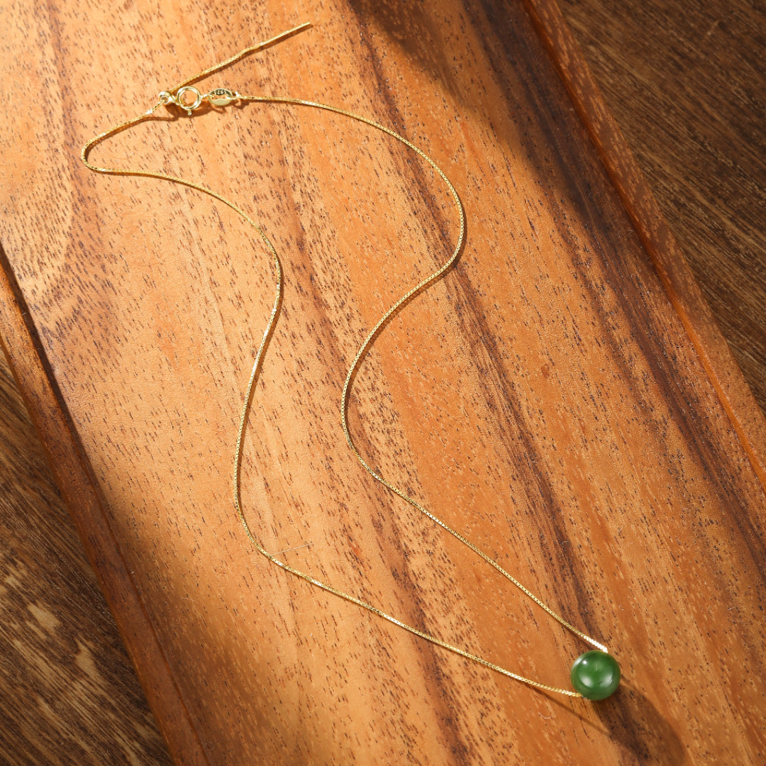 A simple gold chain necklace with a single, round, deep green jade pendant, resting on a wooden surface.