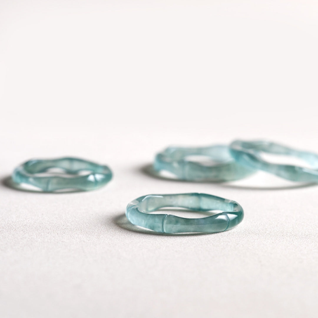 Four translucent blue jade bamboo rings with a wavy, organic shape displayed on a light gray surface against a blurred white background.