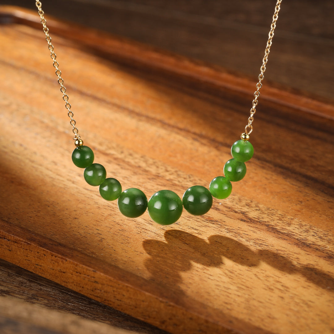 All Smiles Green Jade Beaded Necklace with a delicate gold chain and varying sizes of smooth green jade beads arranged in a gentle curve on a wooden surface.