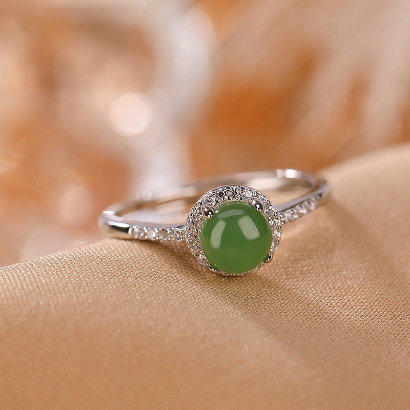 Answered - A round, green jade solitaire gemstone is set in a silver ring with a halo of small, clear stones. The ring is displayed on a soft, textured fabric.