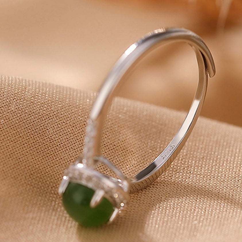 Answered - A delicate silver band ring with a small, round, green jade stone set in a halo of clear cubic zirconia stones, resting on a textured beige fabric surface.