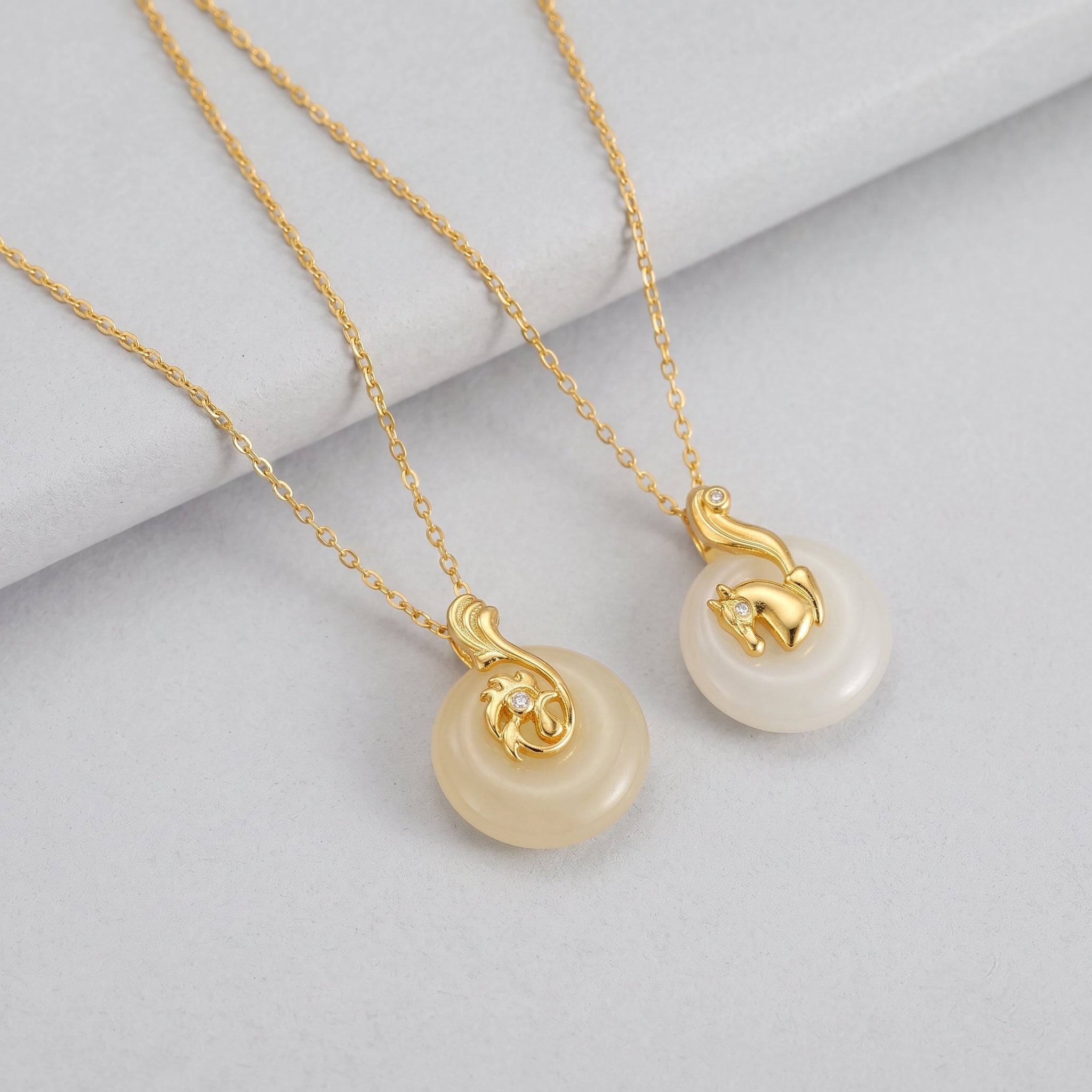 Two Birthright zodiac pendant necklaces with gold chains and white Hetian jade discs, one featuring a gold horse charm with a diamond accent, and the other a gold phoenix-like charm with a diamond accent.