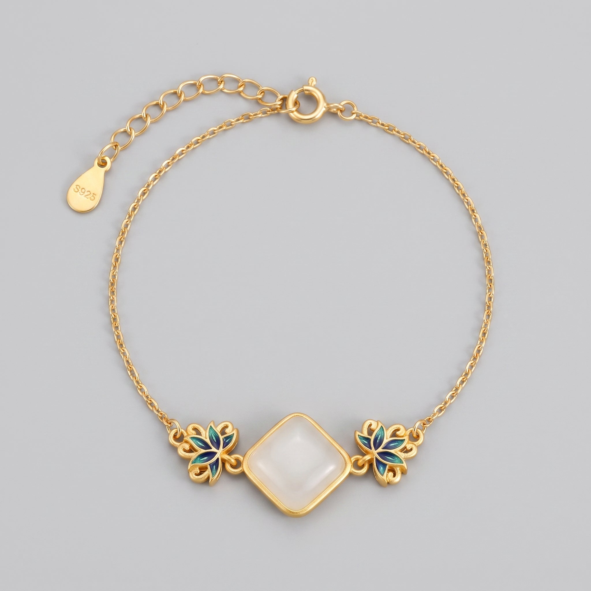 A delicate gold chain bracelet featuring a central square-cut white jade stone, flanked by two ornate blue and green lotus flower charms. The bracelet has an adjustable clasp and a small 'S925' tag.