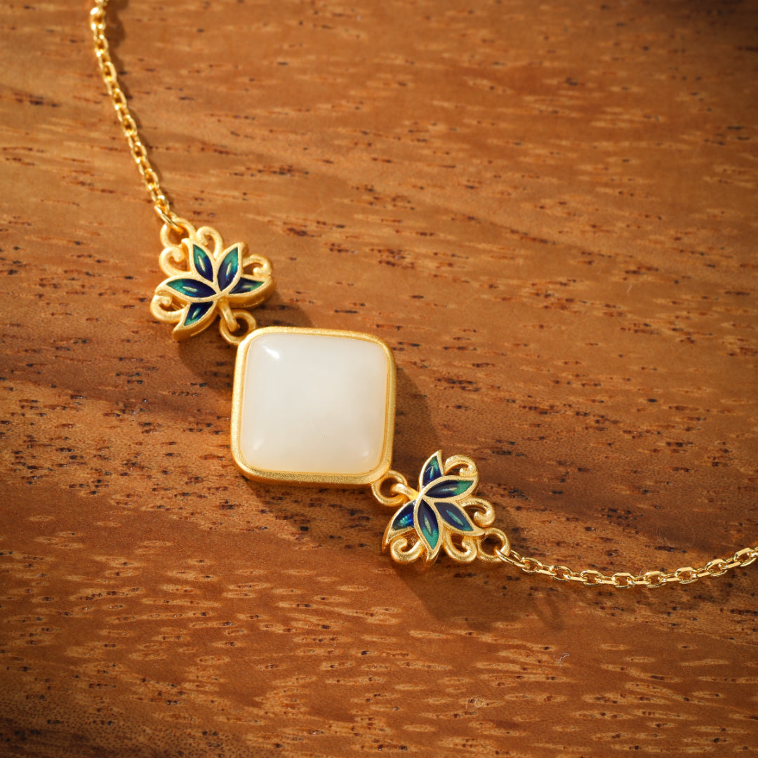 A close-up shot of a gold-plated silver bracelet featuring a square, white Hetian jade stone centerpiece, flanked by two ornate lotus flower charms with blue and green enamel detailing.