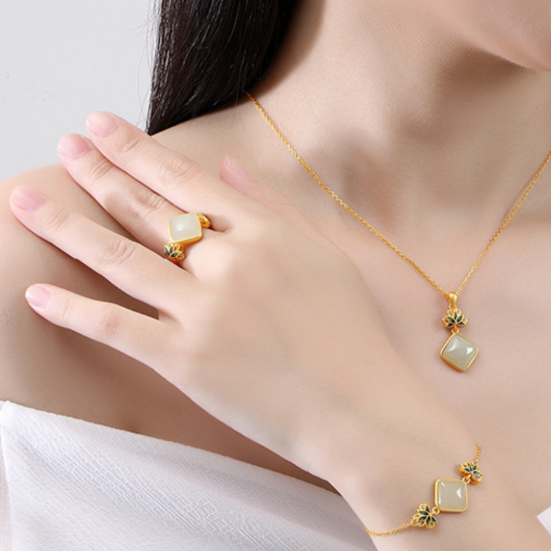 A woman's hand and arm adorned with a gold-plated silver ring and bracelet, both featuring square-shaped white jade stones and delicate blue lotus accents. A matching pendant necklace hangs from her neck.