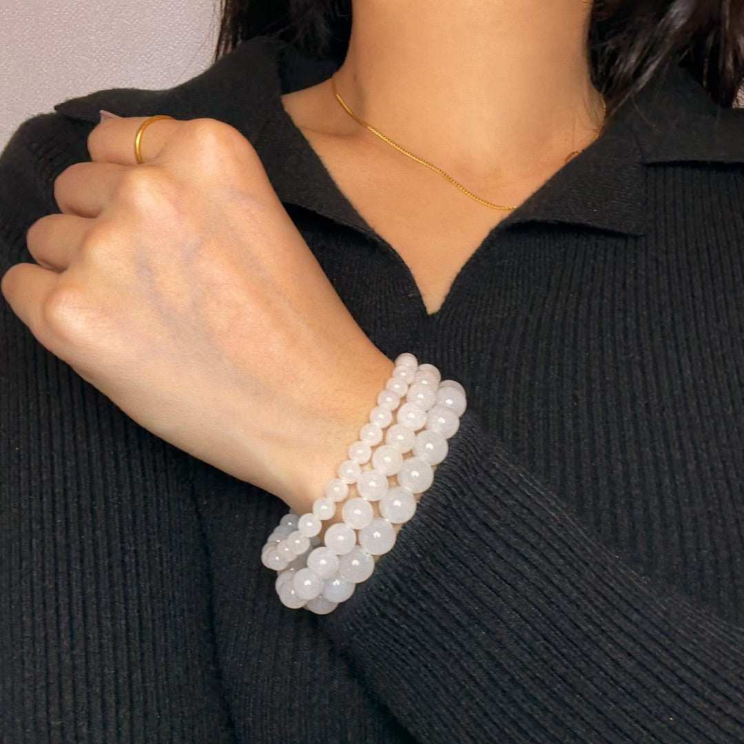 Three stacked bracelets made of smooth, white Hetian jade beads in varying sizes, worn on a woman's wrist over a black sweater.