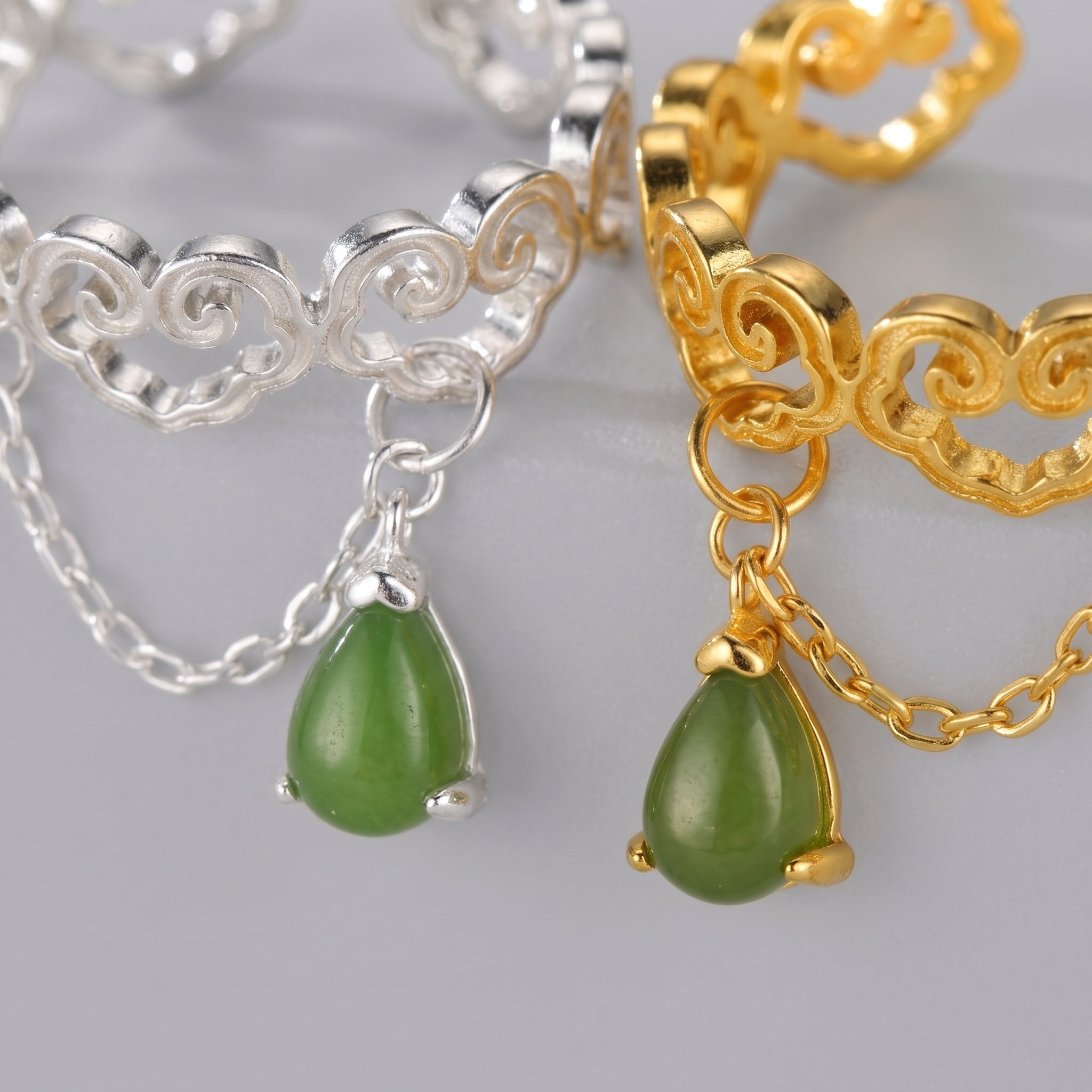 Close-up of two cloud-shaped rings, one silver and one gold-plated, each with a dangling teardrop-shaped green jade gemstone and a delicate chain.