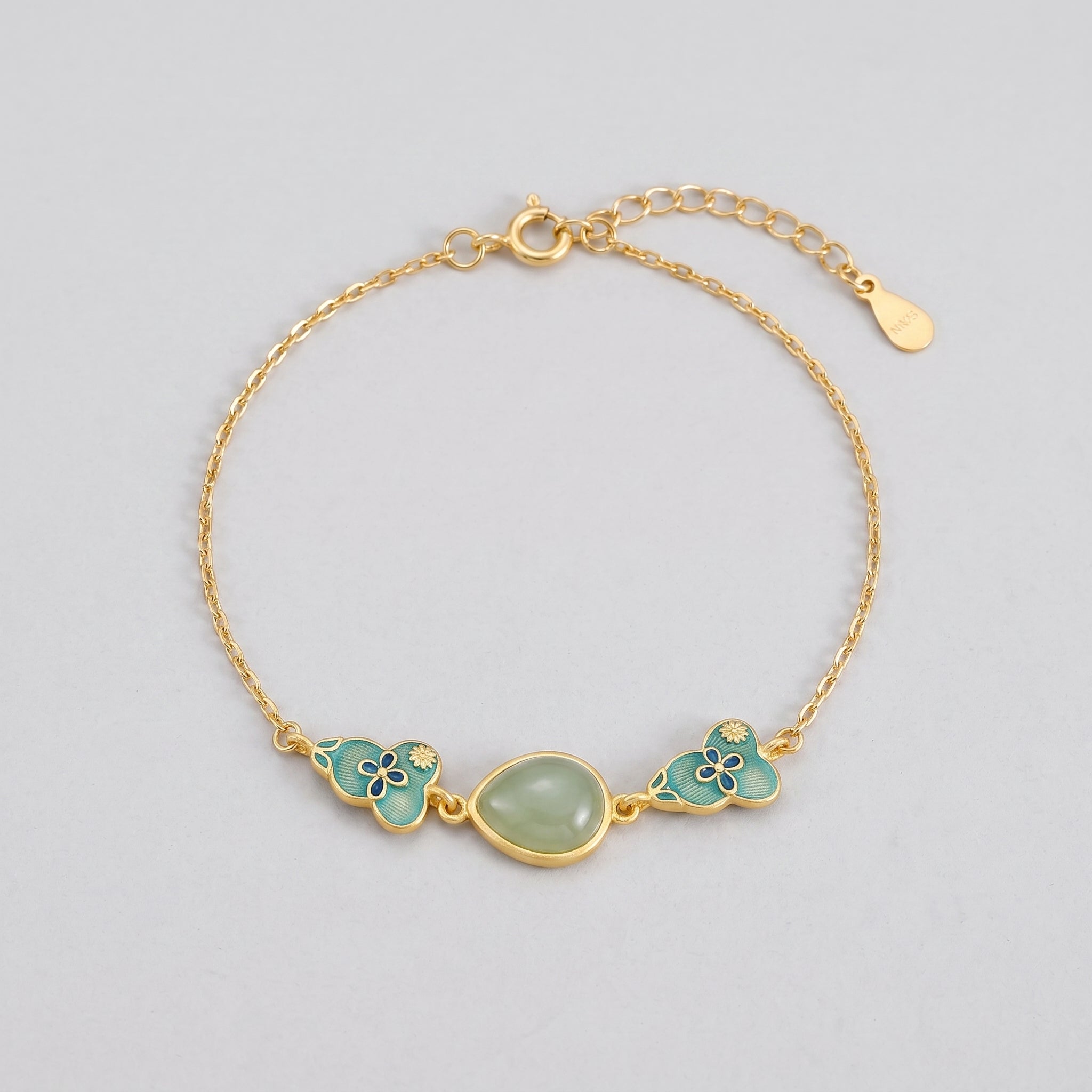 Gold plated silver bracelet with a central oval light green jade stone, flanked by two teal cloud-shaped charms with blue floral patterns.