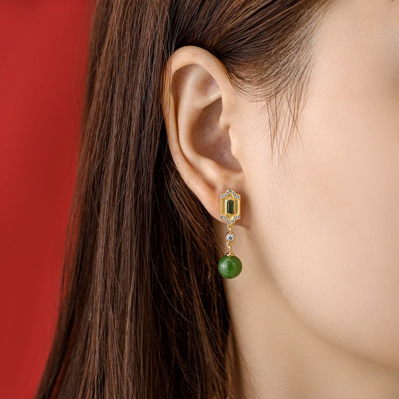 Close-up of a woman wearing Kindred green jade and emerald drop earrings, featuring a gold setting with small diamonds and a dangling green jade bead.