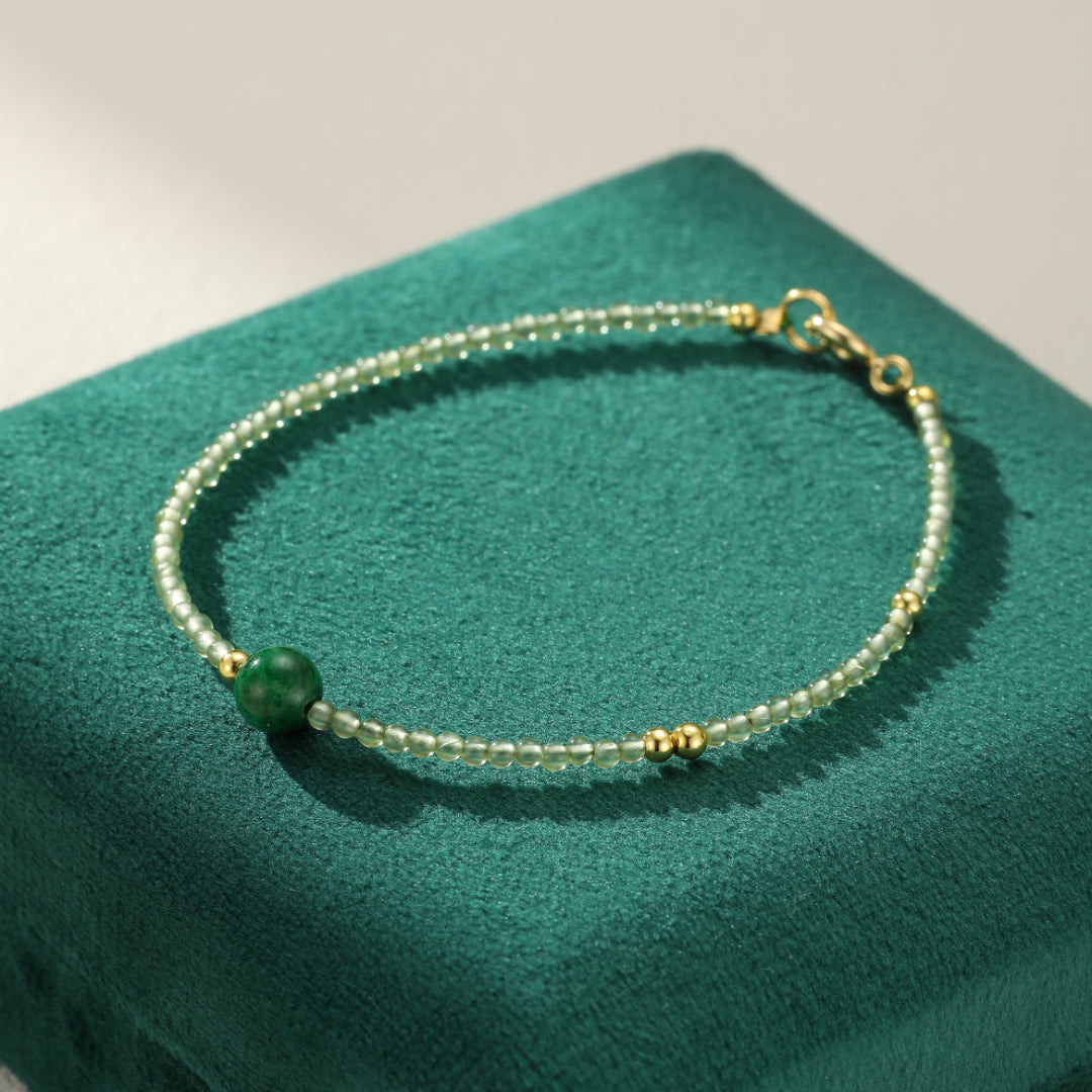 A delicate bracelet made of small, light green beads and gold accents, featuring a single, larger dark green jade bead. The bracelet is displayed on a textured dark green velvet surface.