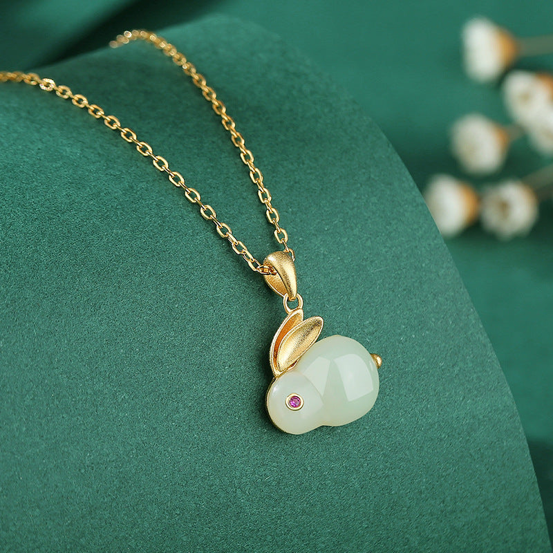 Moonglow Hetian Jade Rabbit Pendant Necklace with a gold chain and a small pink gemstone eye, set against a dark green background with blurred white flowers.