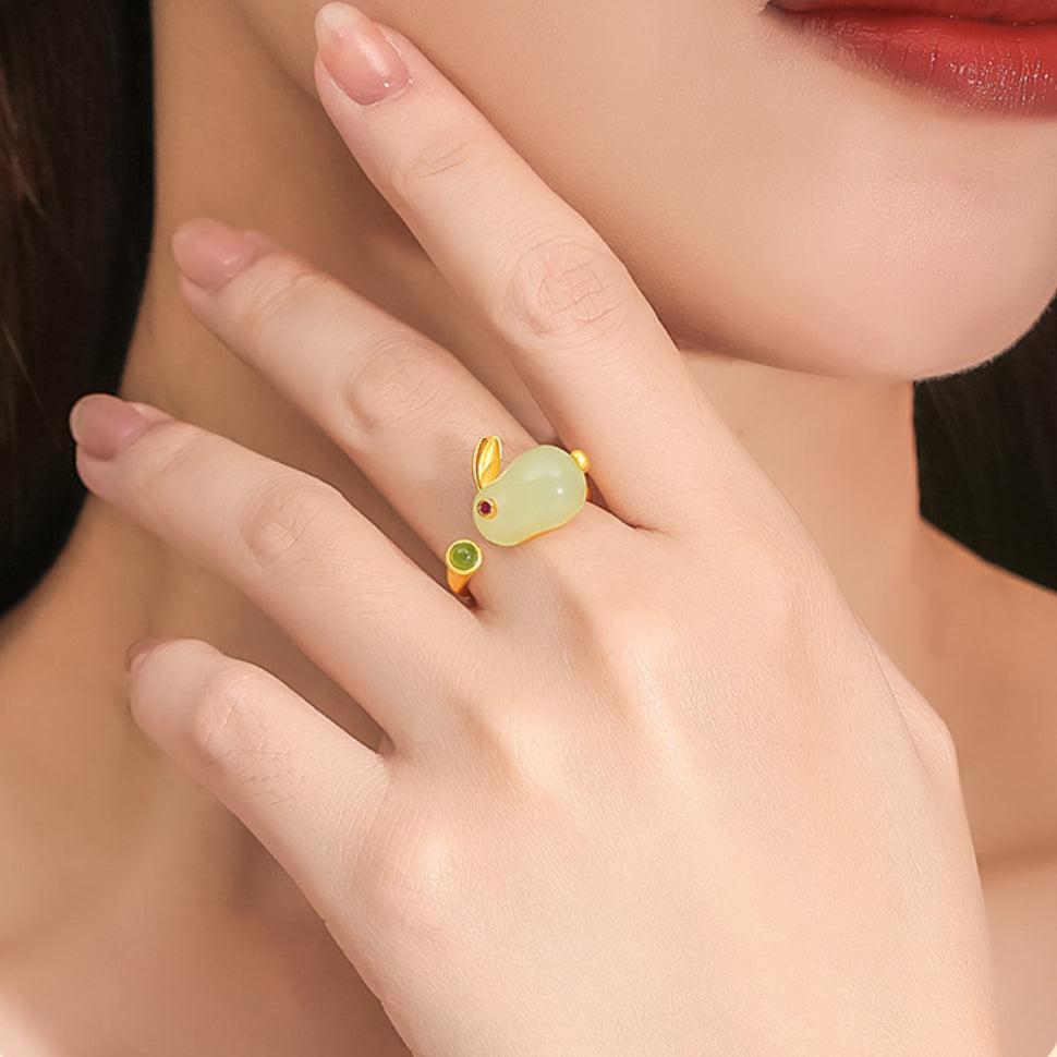 Close-up of a woman's hand wearing the Moonglow Hetian Jade Rabbit Ring, featuring a gold-plated silver band and a carved jade rabbit with red eyes and a green gemstone accent.