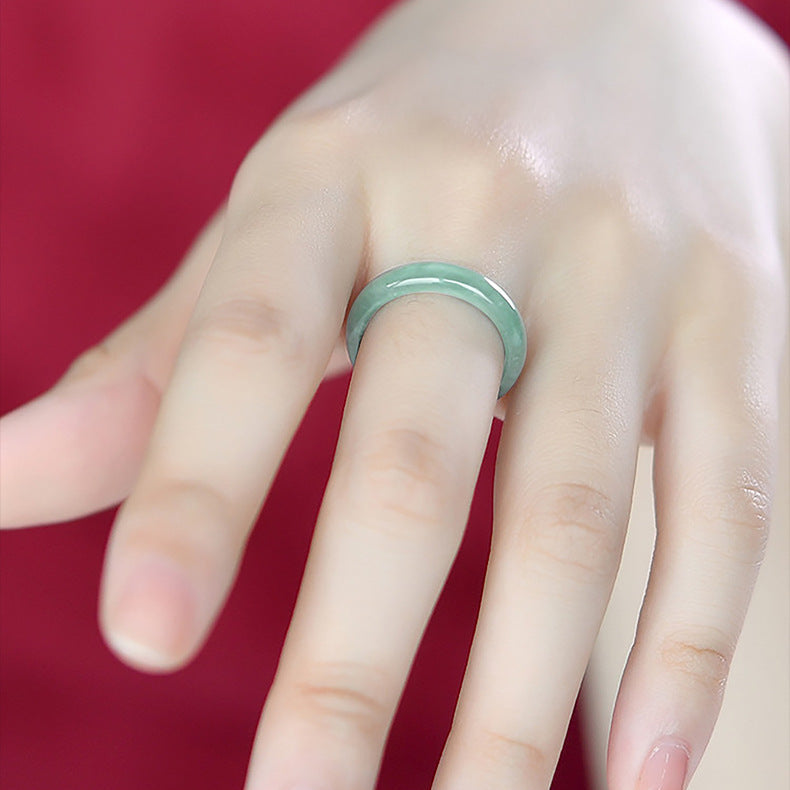 A close-up shot of a hand wearing the Same Sky Ice Jade Couple Band Ring on the ring finger. The ring is a simple, smooth band made of translucent pale green jade, showcasing its natural beauty and delicate icy luster against a deep red background.