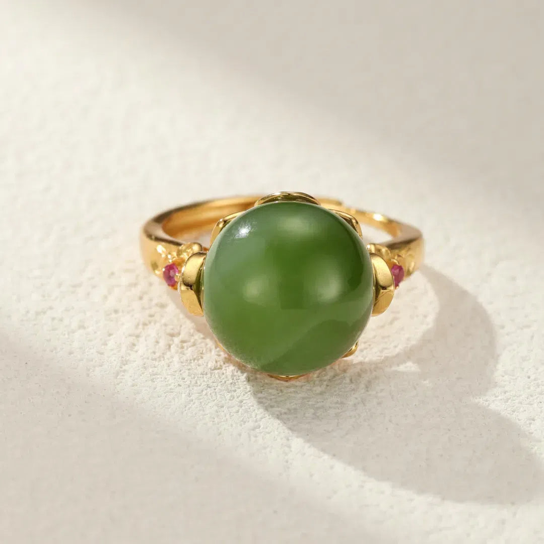 A gold-plated silver ring featuring a large, round, smooth green jade stone as the centerpiece, with small red accent stones on either side of the jade.