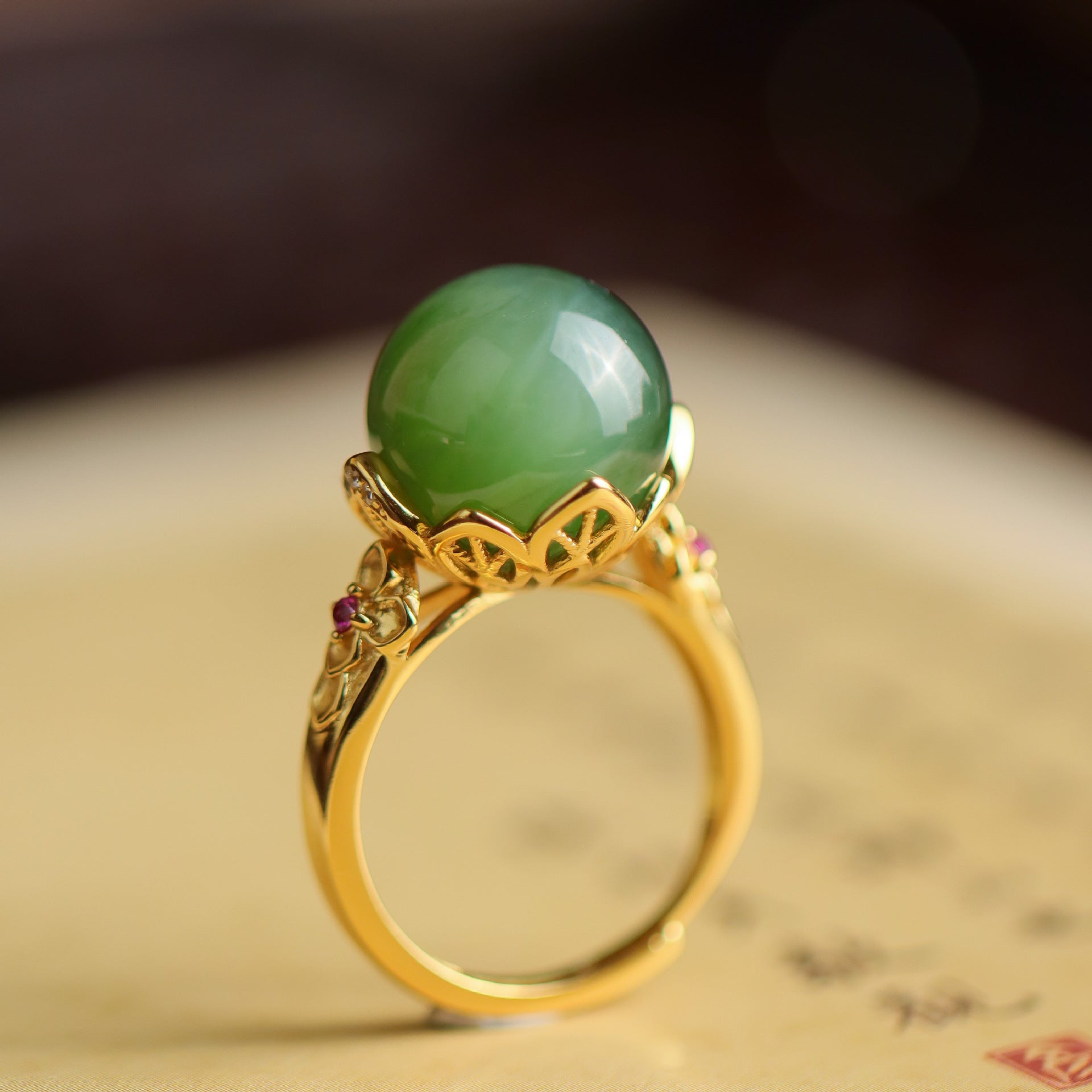 A detailed close-up of the Sovereign ring, showcasing a smooth, round, vibrant green jade gemstone set in an ornate gold-toned metal band with floral motifs and small ruby accents.