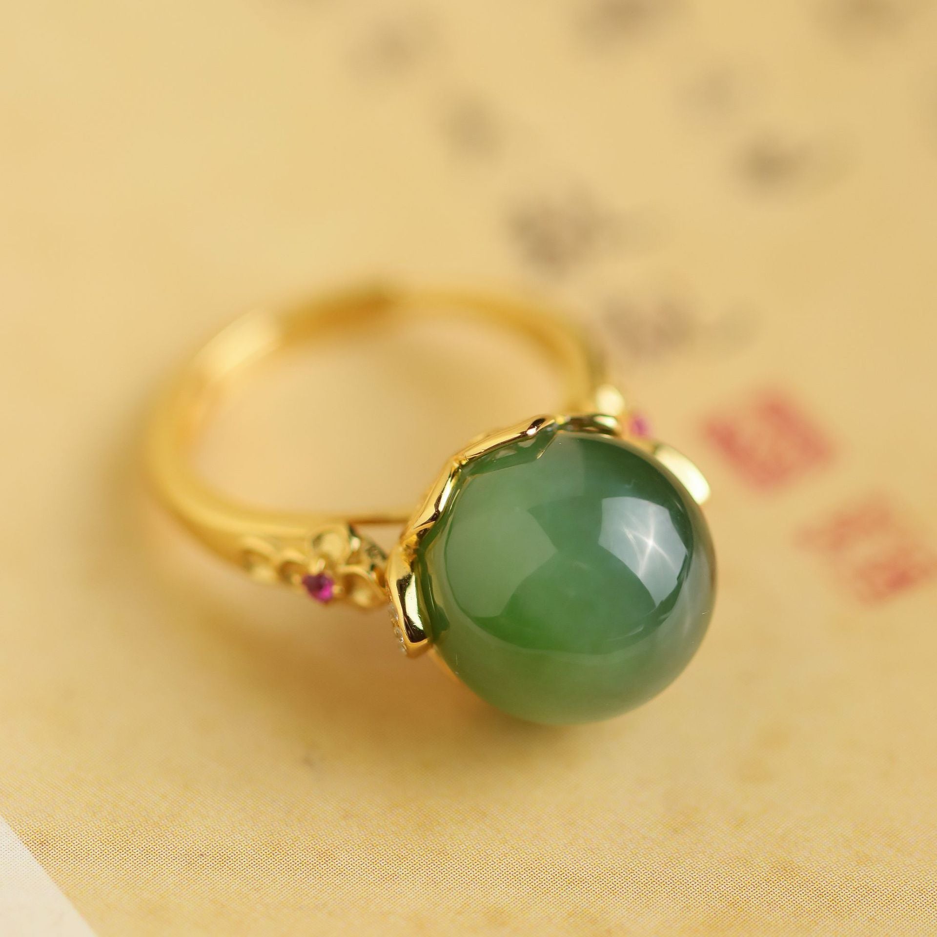 A round, green jade solitaire gemstone is set in a gold-toned ring with small ruby accents on the band.
