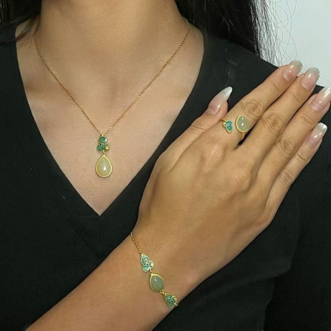 A woman models a jewelry set featuring a gold necklace with a teardrop jade pendant, a matching ring with a green jade stone, and a bracelet with jade beads and green accents.